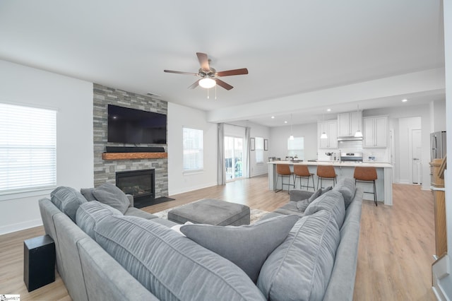 living area with light wood-type flooring, recessed lighting, a stone fireplace, baseboards, and ceiling fan