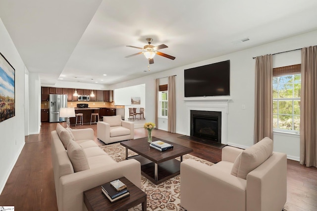 living area with a glass covered fireplace, baseboards, dark wood-style floors, visible vents, and ceiling fan