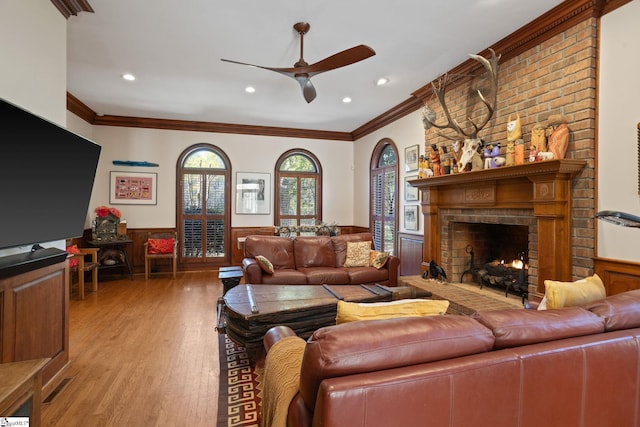 living area with crown molding, a ceiling fan, wood finished floors, a brick fireplace, and wainscoting