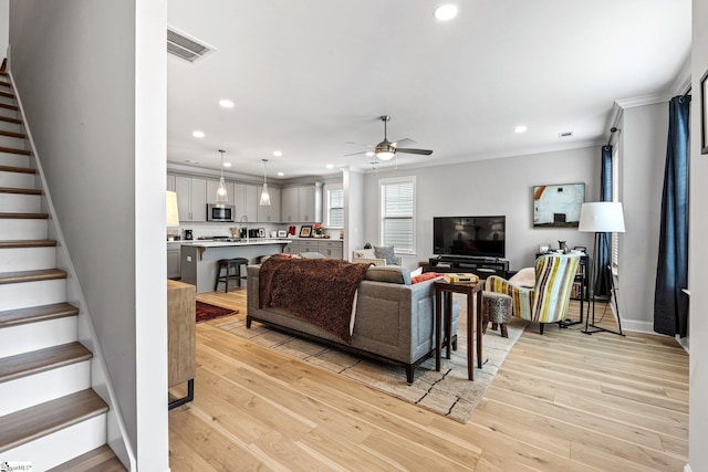 living area featuring stairway, ornamental molding, ceiling fan, light wood-style flooring, and visible vents