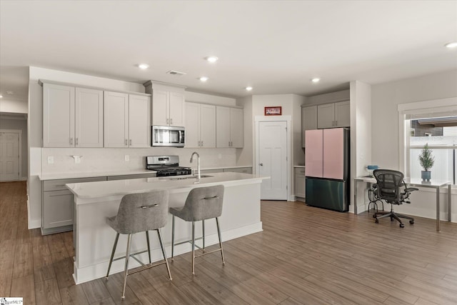 kitchen featuring a sink, backsplash, wood finished floors, appliances with stainless steel finishes, and a kitchen island with sink