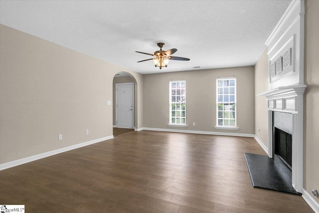 unfurnished living room featuring dark wood-style floors, arched walkways, a fireplace with raised hearth, and ceiling fan