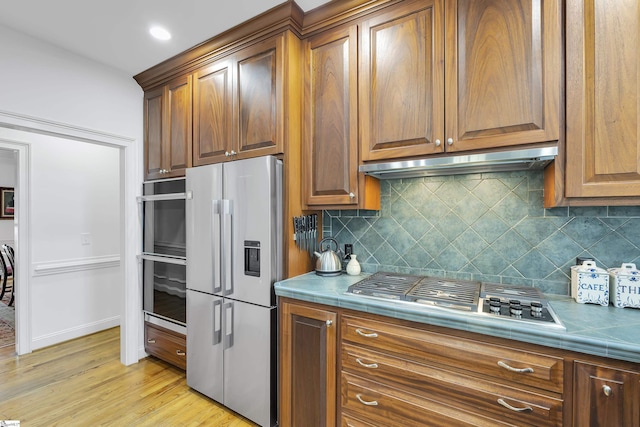 kitchen featuring stainless steel appliances, under cabinet range hood, decorative backsplash, tile countertops, and light wood-style flooring