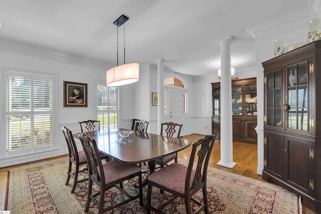 dining area featuring crown molding, ornate columns, a decorative wall, light wood-style flooring, and wainscoting