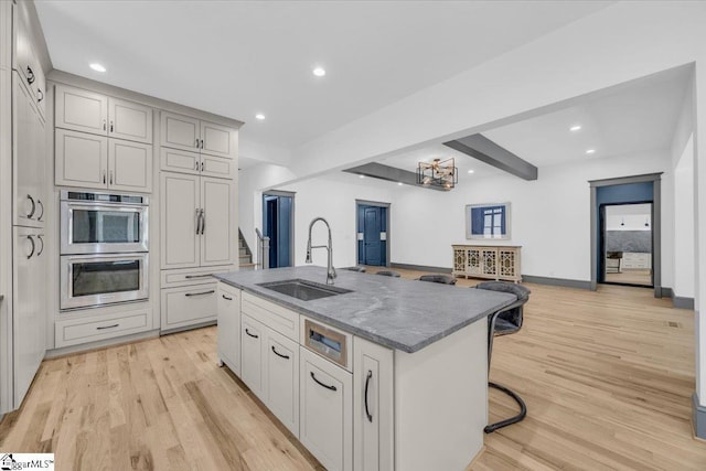 kitchen with a sink, a kitchen island with sink, stainless steel double oven, and light wood finished floors