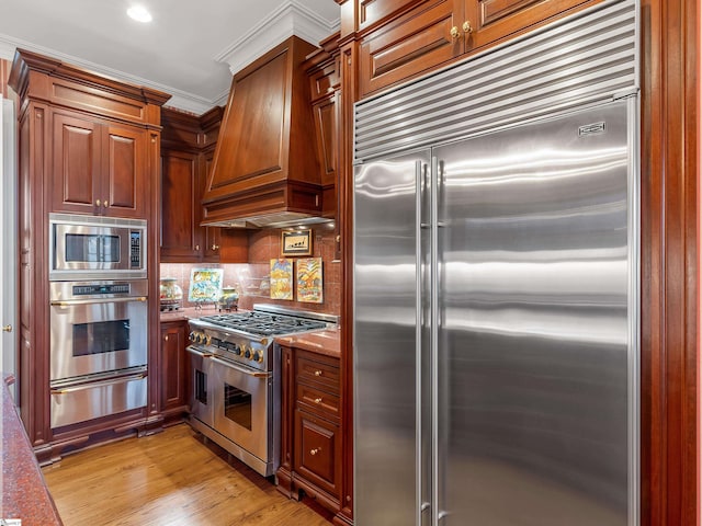 kitchen featuring a warming drawer, light wood-type flooring, premium range hood, ornamental molding, and built in appliances