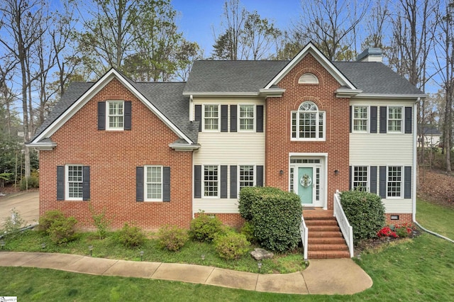 colonial home with a chimney, a front yard, brick siding, and roof with shingles