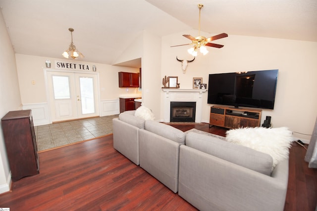 living area featuring a fireplace, vaulted ceiling, ceiling fan with notable chandelier, dark wood-type flooring, and a wainscoted wall