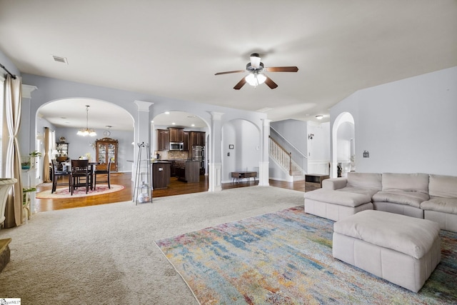 carpeted living room featuring stairway, visible vents, ornate columns, arched walkways, and ceiling fan with notable chandelier