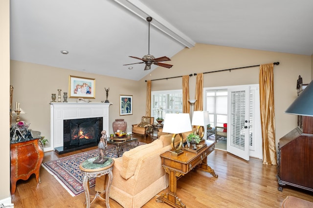 living area featuring lofted ceiling with beams, wood finished floors, a warm lit fireplace, and ceiling fan