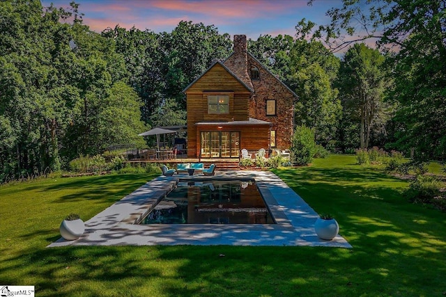back of house at dusk with a view of trees, a yard, a wooden deck, and a chimney