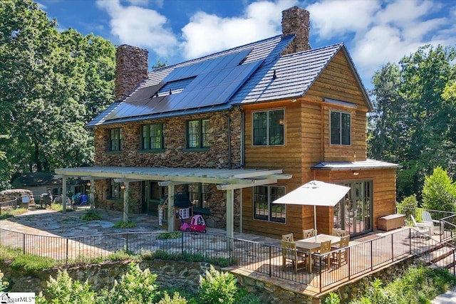 rear view of house featuring solar panels, stone siding, a chimney, fence, and a patio