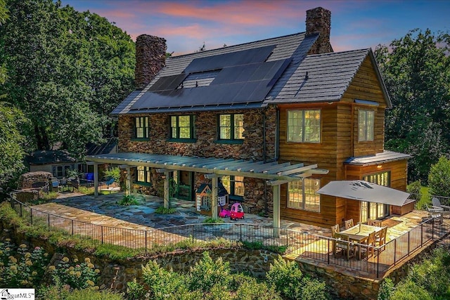 rear view of property featuring a patio area, solar panels, stone siding, and a chimney