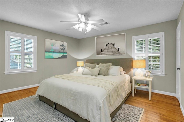 bedroom with ceiling fan, a textured ceiling, visible vents, wood finished floors, and baseboards