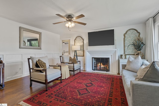 living area with a decorative wall, a lit fireplace, a ceiling fan, a wainscoted wall, and wood finished floors