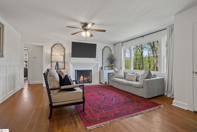 living area featuring a decorative wall, ceiling fan, a fireplace with flush hearth, a wainscoted wall, and wood-type flooring