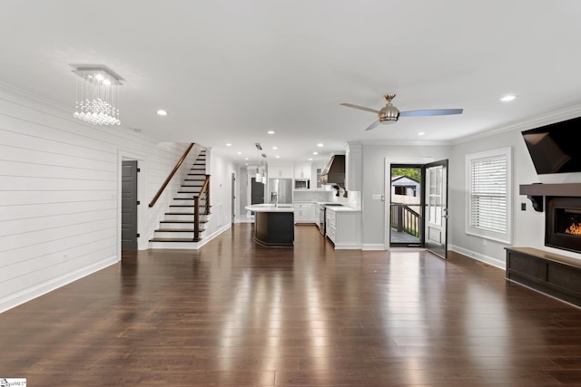 unfurnished living room with baseboards, dark wood finished floors, crown molding, ceiling fan with notable chandelier, and a glass covered fireplace