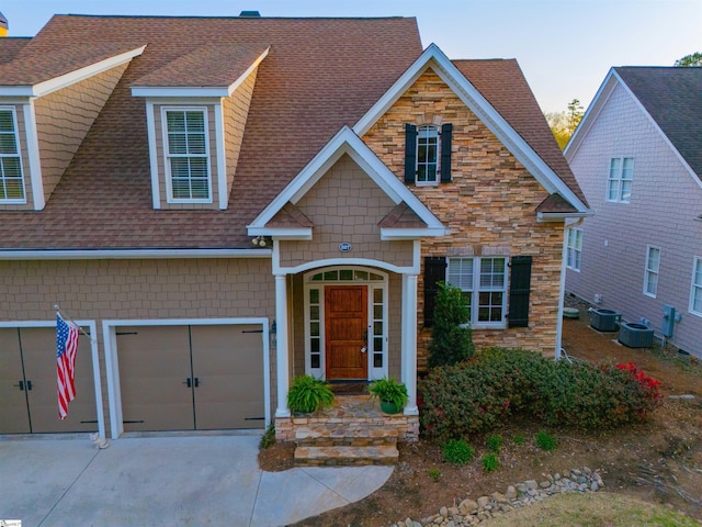 view of front of house featuring central AC unit, an attached garage, stone siding, driveway, and roof with shingles