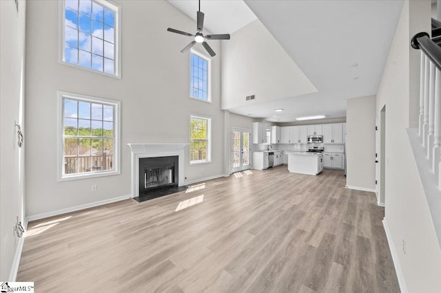 unfurnished living room featuring baseboards, ceiling fan, light wood-style floors, a fireplace, and a towering ceiling