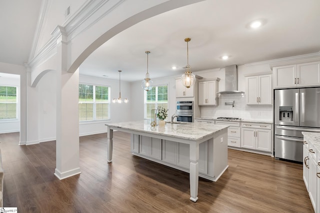 kitchen featuring light stone countertops, a center island with sink, wall chimney exhaust hood, stainless steel appliances, and arched walkways