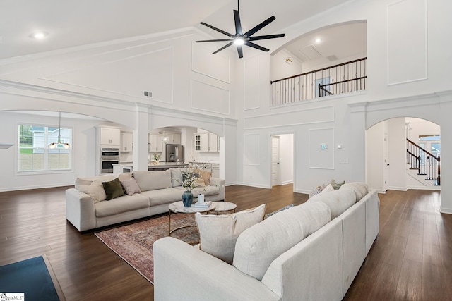 living room with a towering ceiling, stairway, dark wood-style floors, a ceiling fan, and arched walkways