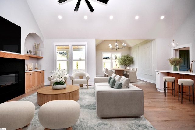 living room featuring a glass covered fireplace, light wood-type flooring, baseboards, recessed lighting, and ceiling fan with notable chandelier