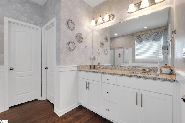 bathroom with wood finished floors, a sink, a stall shower, wallpapered walls, and double vanity