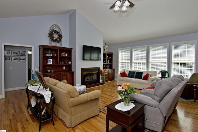 living room with high vaulted ceiling, ceiling fan, a glass covered fireplace, light wood finished floors, and baseboards