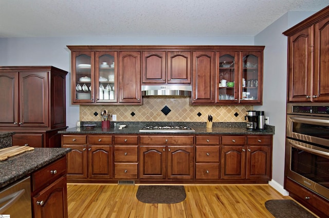 kitchen with glass insert cabinets, wall chimney range hood, light wood-type flooring, appliances with stainless steel finishes, and decorative backsplash