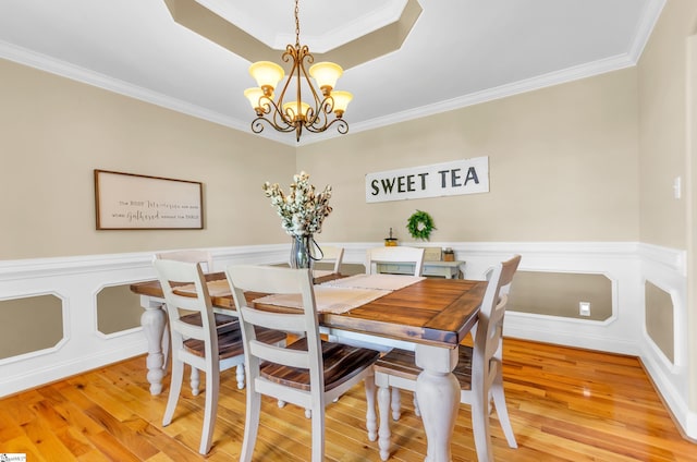 dining room with a chandelier, ornamental molding, light wood-style floors, a raised ceiling, and wainscoting