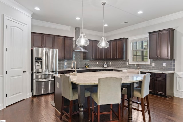 kitchen featuring light stone counters, a center island with sink, dark brown cabinetry, decorative backsplash, and stainless steel refrigerator with ice dispenser