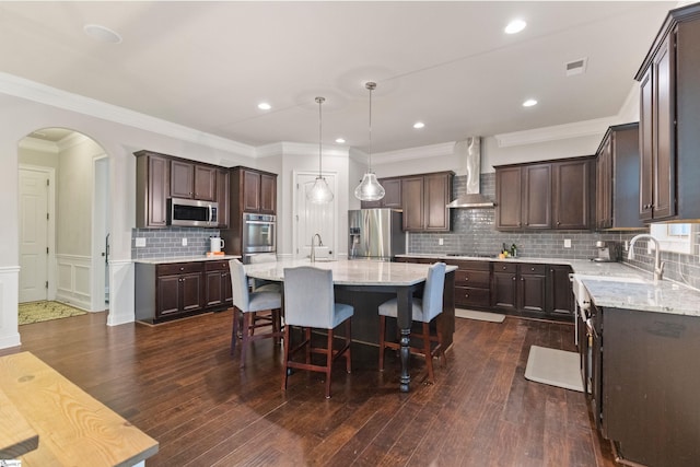 kitchen with wall chimney exhaust hood, arched walkways, dark brown cabinets, and appliances with stainless steel finishes