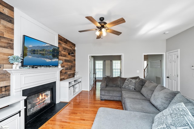 living room with light wood-style floors, a glass covered fireplace, wooden walls, and ceiling fan