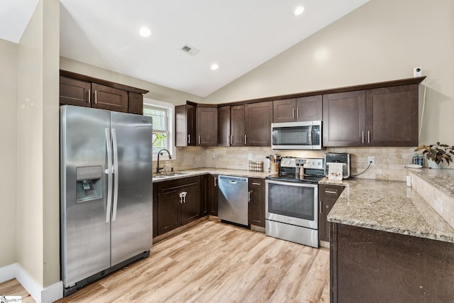 kitchen with a sink, dark brown cabinets, vaulted ceiling, light stone countertops, and appliances with stainless steel finishes