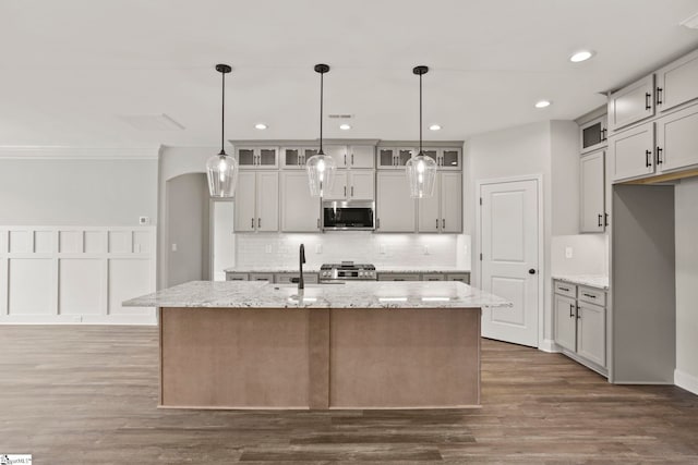 kitchen with dark wood-style floors, backsplash, stainless steel microwave, a center island with sink, and glass insert cabinets