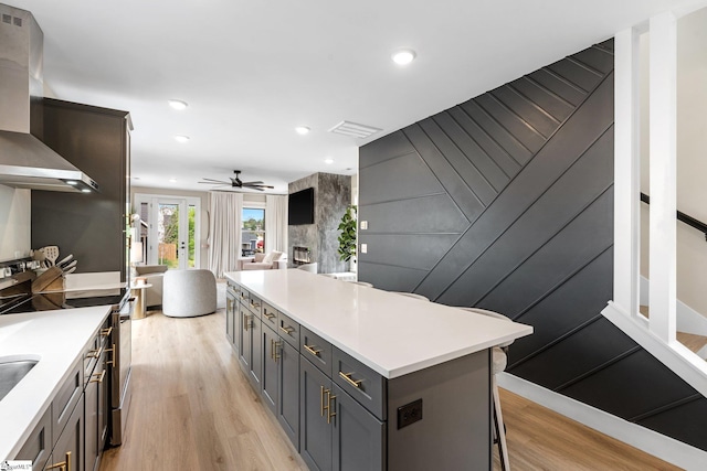 kitchen featuring light wood-type flooring, a ceiling fan, wall chimney range hood, light countertops, and a kitchen island