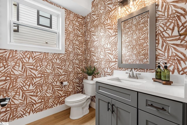 half bath with wallpapered walls, wood finished floors, toilet, and vanity