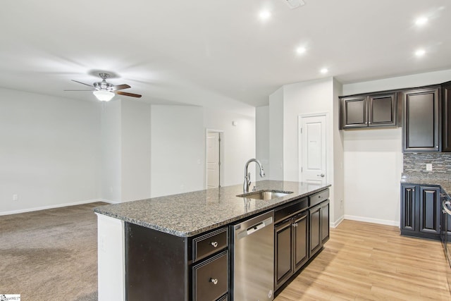 kitchen featuring stainless steel dishwasher, a kitchen island with sink, dark stone counters, a sink, and baseboards