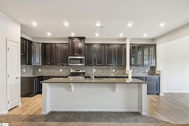 kitchen featuring dark stone counters, tasteful backsplash, a sink, stainless steel appliances, and a kitchen island with sink