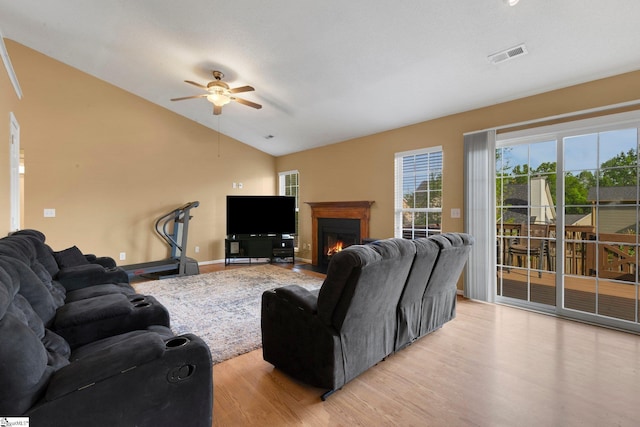 living area featuring light wood finished floors, vaulted ceiling, visible vents, a ceiling fan, and a lit fireplace