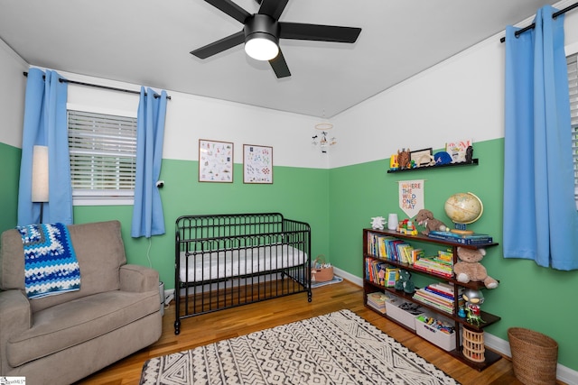 bedroom featuring ceiling fan, a nursery area, wood finished floors, and baseboards