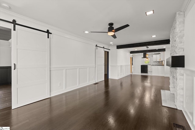 unfurnished living room with a barn door, a ceiling fan, dark wood-type flooring, and a decorative wall