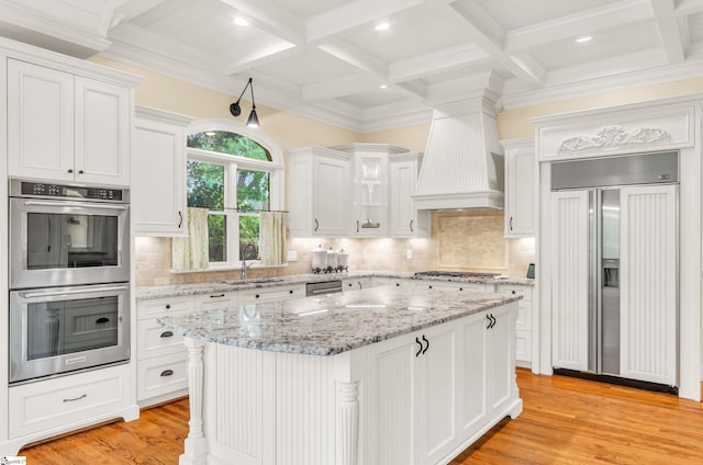 kitchen with appliances with stainless steel finishes, a sink, premium range hood, white cabinetry, and coffered ceiling