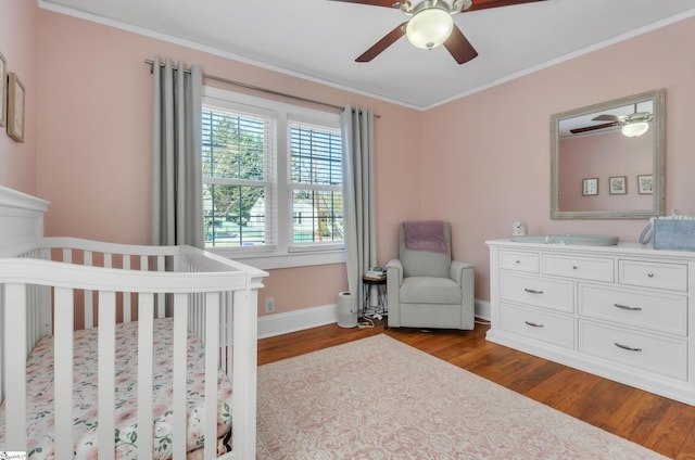 bedroom with crown molding, baseboards, dark wood-style floors, a crib, and a ceiling fan