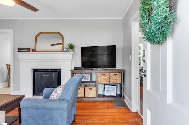 living area featuring ornamental molding, a brick fireplace, wood finished floors, baseboards, and ceiling fan