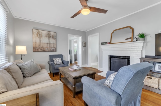 living area featuring a brick fireplace, ceiling fan, crown molding, and wood finished floors