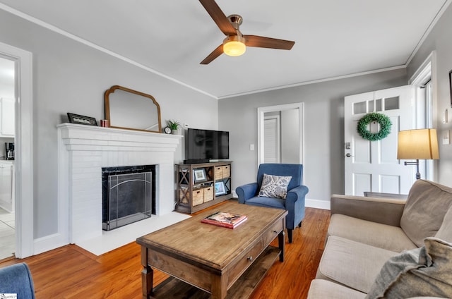 living room with crown molding, a brick fireplace, wood finished floors, baseboards, and a ceiling fan
