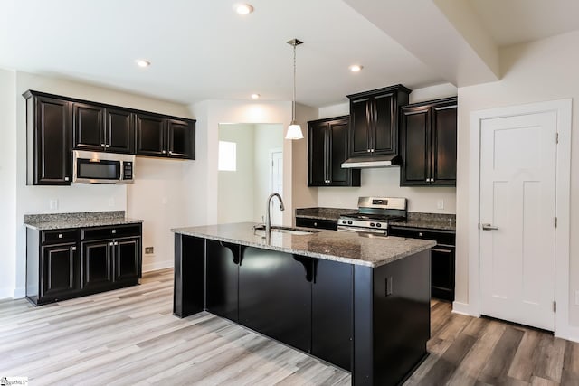 kitchen with appliances with stainless steel finishes, a sink, dark stone countertops, dark cabinets, and light wood-style flooring