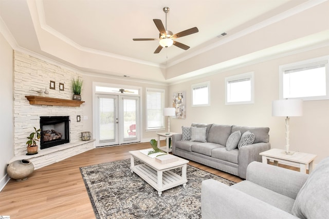 living area featuring a raised ceiling, light wood finished floors, a stone fireplace, a ceiling fan, and ornamental molding