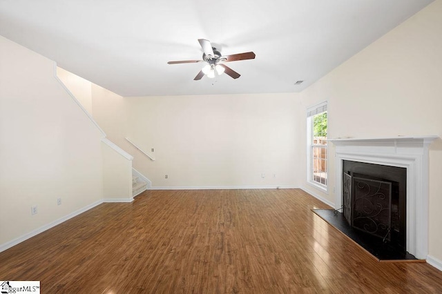 unfurnished living room with ceiling fan, a fireplace with raised hearth, baseboards, wood finished floors, and stairway